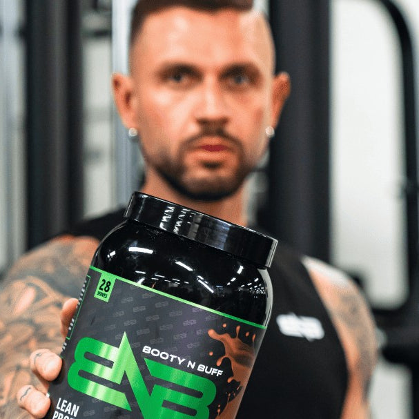 A man in the gym holding out a tub of Booty N Buff Lean Protein, Dark Chocolate Coconut.