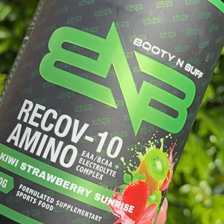 A hand holding a tub of Booty N Buff Recov-10 Amino, Kiwi Strawberry Sunrise, outdoors.