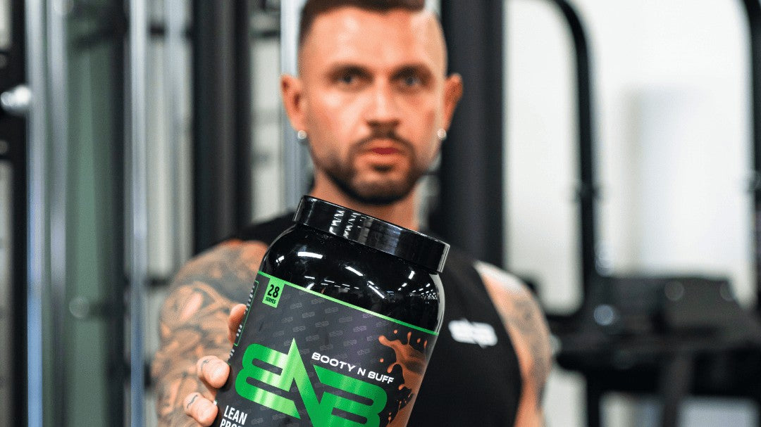 A man in the gym holding out a tub of Booty N Buff Lean Protein, Dark Chocolate Coconut.