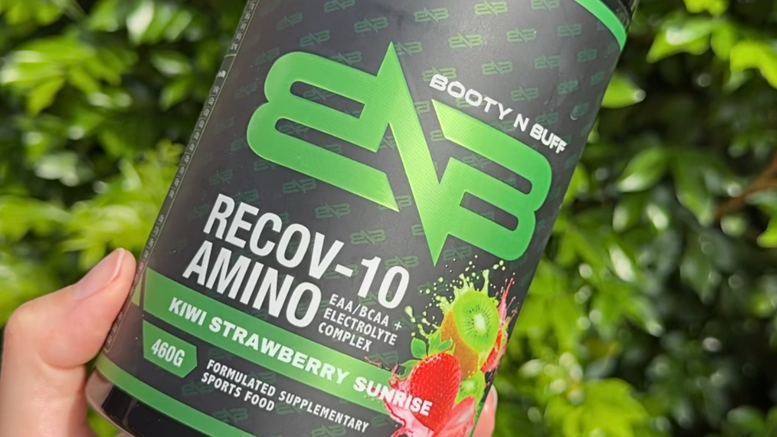 A hand holding a tub of Booty N Buff Recov-10 Amino, Kiwi Strawberry Sunrise, outdoors.